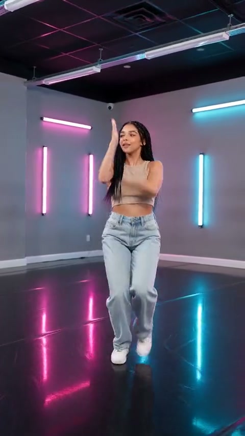 Quadro de um vídeo externo com uma pessoa de jeans e tênis brancos levantando um braço e um pé em um estúdio neon rosa e ciano